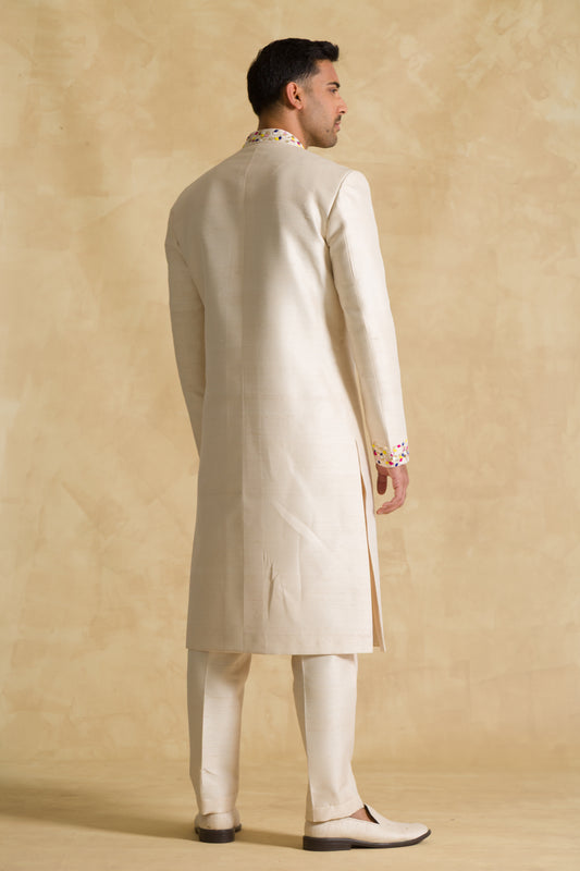 Man in cream sherwani suit with colorful collar, standing against beige background
