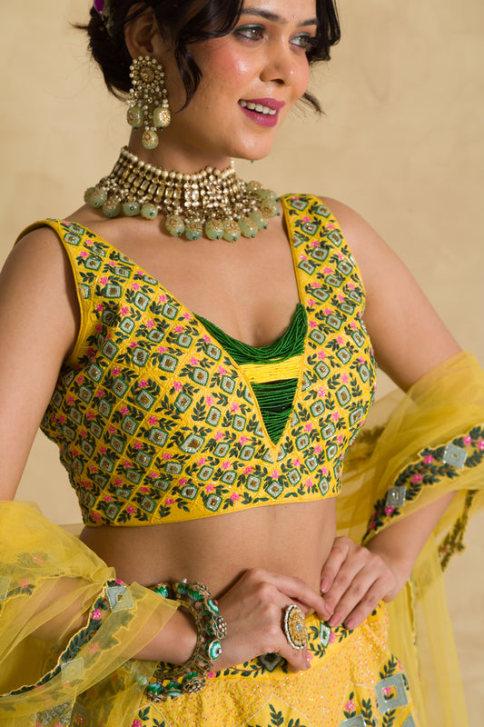 Woman in vibrant yellow embroidered lehenga choli with green jewelry, posing indoors