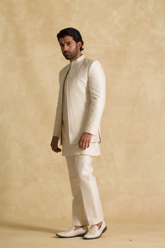 Man wearing an embroidered ivory sherwani suit with matching pants and loafers, beige background