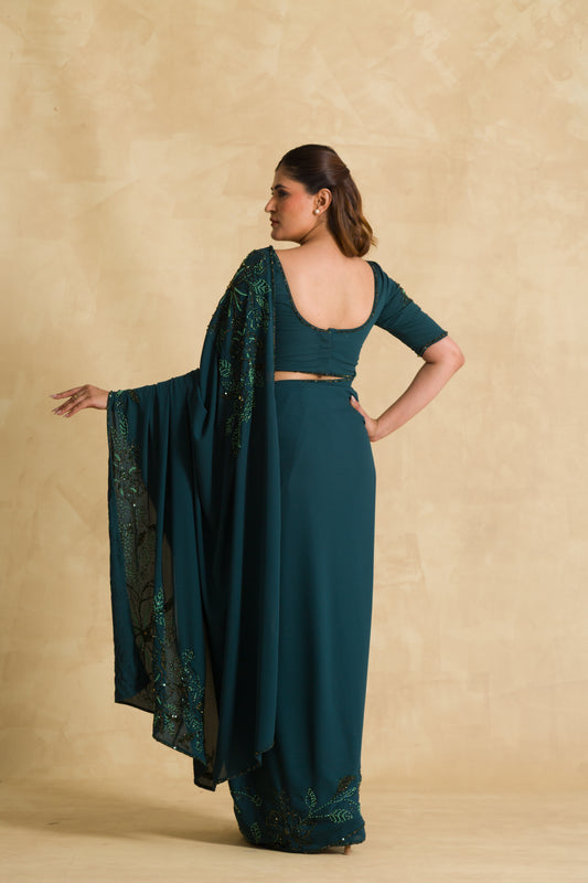 Woman in teal saree with embroidered details, standing against beige background